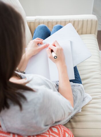 A person with dark shoulder length hair is sitting with their back turned away from the camera. They are writing in a blank journal. 