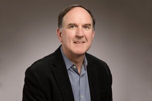 Professor Wood smiles in front of a gray gradient background. He is wearing a black jacket with a blue and white thinly-checkered collared shirt. 