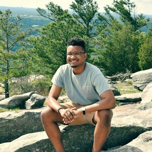 Hollis smiles sitting on a rock in front of trees.