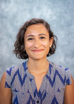 María smiles in front of a grey-blue background. María has short curly hair and is wearing a blue and white chevron blouse.