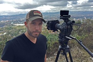 Ryan smiles standing next to a camera on a tripod. He is wearing a hat and a black v-neck shirt.