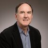 Professor Wood smiles in front of a gray gradient background. He is wearing a black jacket with a blue and white thinly-checkered collared shirt. 