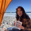 Hilary smiles at the beach. She is holding a book titled Heads of the Colored People.